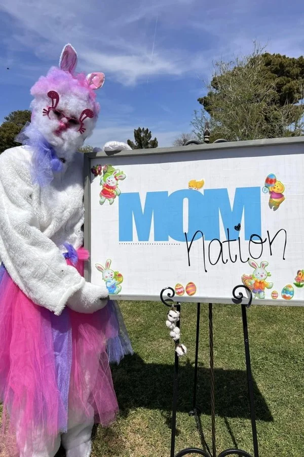 Person dressed in a pink bunny costume with glasses and bunny ears, standing outdoors next to a sign that says "MOM" with added words "nation" and decorated with Easter-themed stickers.