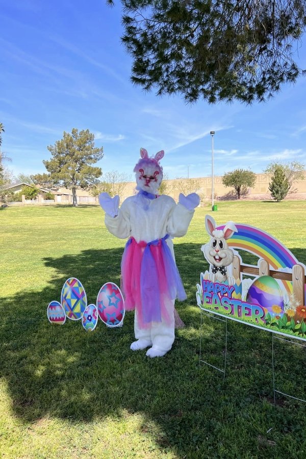 Easter-themed outdoor scene with a person in a bunny costume, Bunny Ears, standing on grass near colorful decorated Easter eggs, a rainbow, and a sign that reads 'Happy Easter' featuring a smiling cartoon bunny.