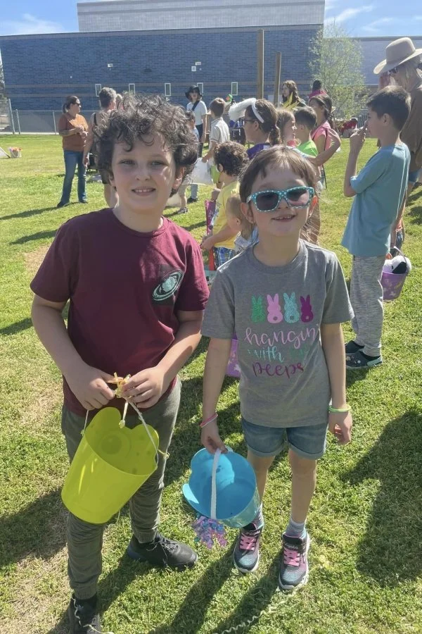 Two children, a boy and a girl, standing on grass during an Easter egg hunt, holding yellow and blue baskets, with other kids and adults in the background.