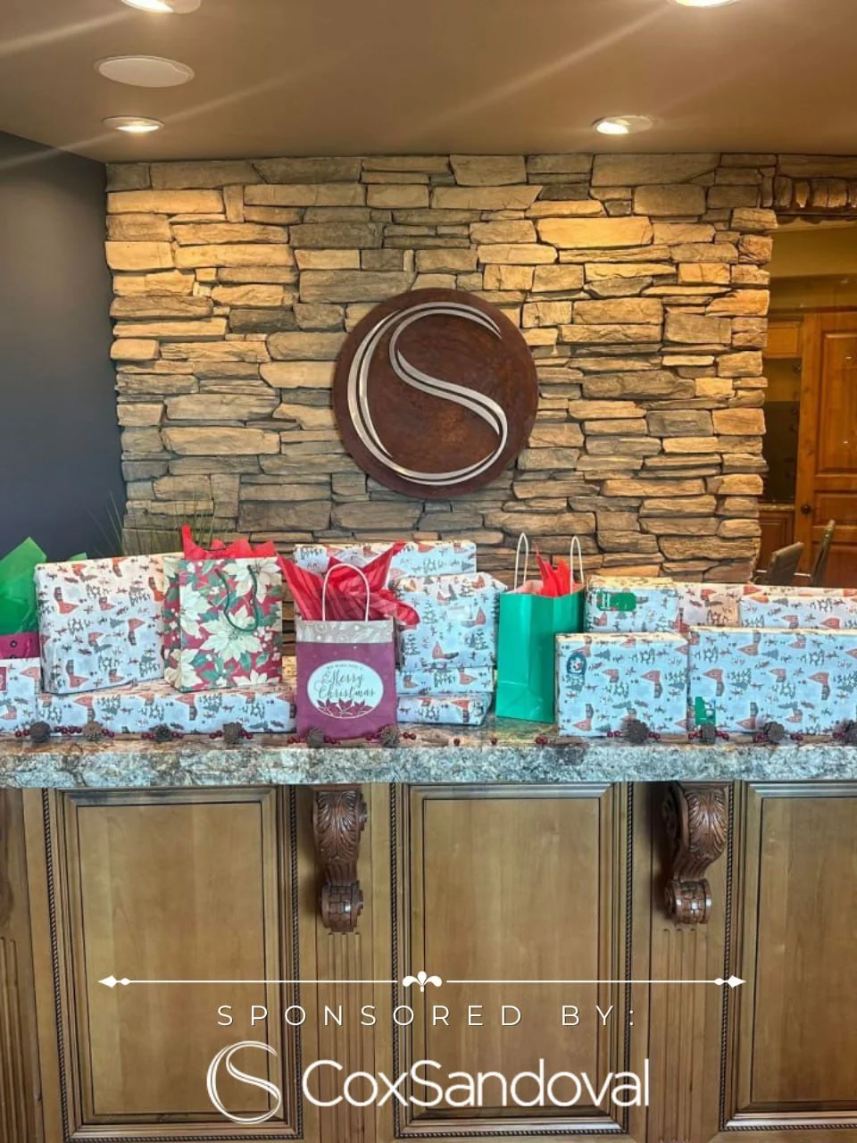 Christmas gifts wrapped in festive paper and gift bags, placed on a granite countertop in front of a stone wall with a wooden decorative piece showing the letter 'S'.