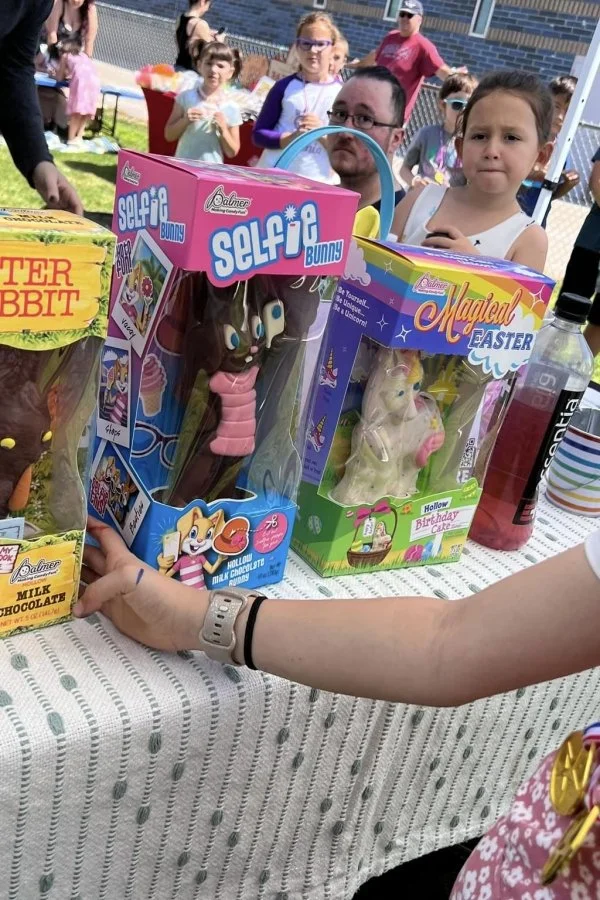 Children at an outdoor birthday party with various chocolate Easter bunnies and toys on a table, some children holding snacks, and a woman’s arm in the foreground.
