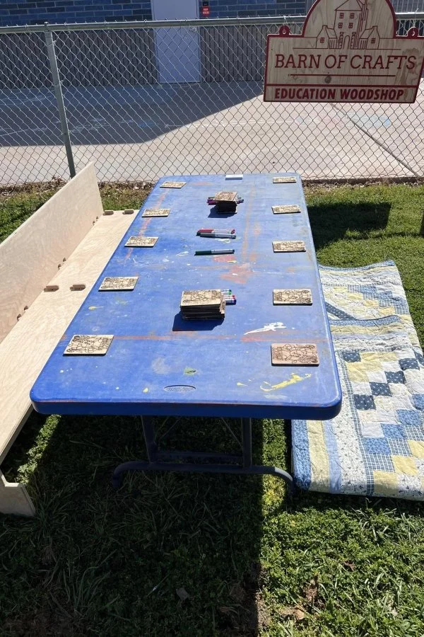 A blue table with wooden tiles and small colored markers on it, outdoors on grass, with a fenced area and a sign that reads 'Barn of Crafts Education Woodshop.'