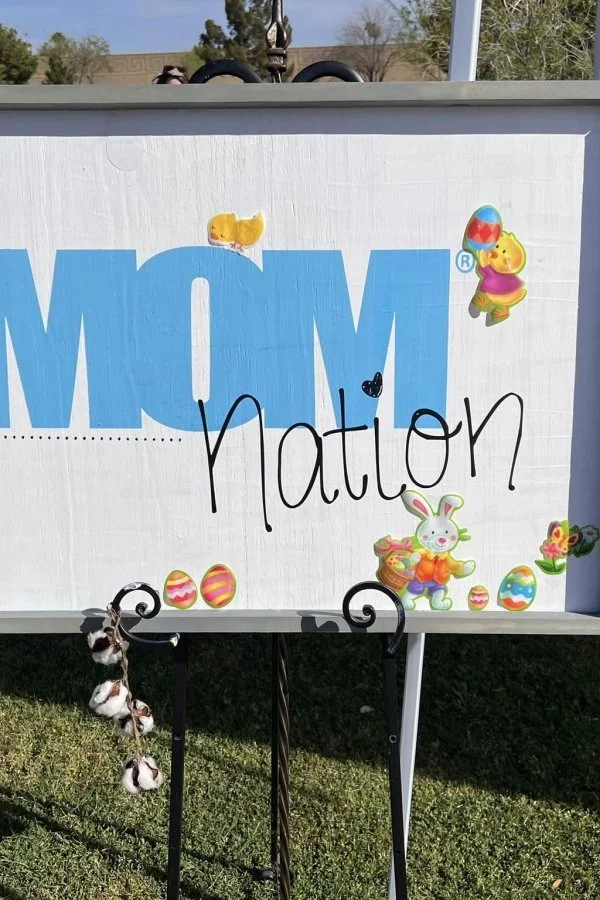 Easter-themed sign with large blue letters spelling 'AMOM' and the word 'nation' handwritten over it; decorated with cartoon stickers of bunnies, chicks, Easter eggs, and flowers, placed outdoors on a grassy surface.