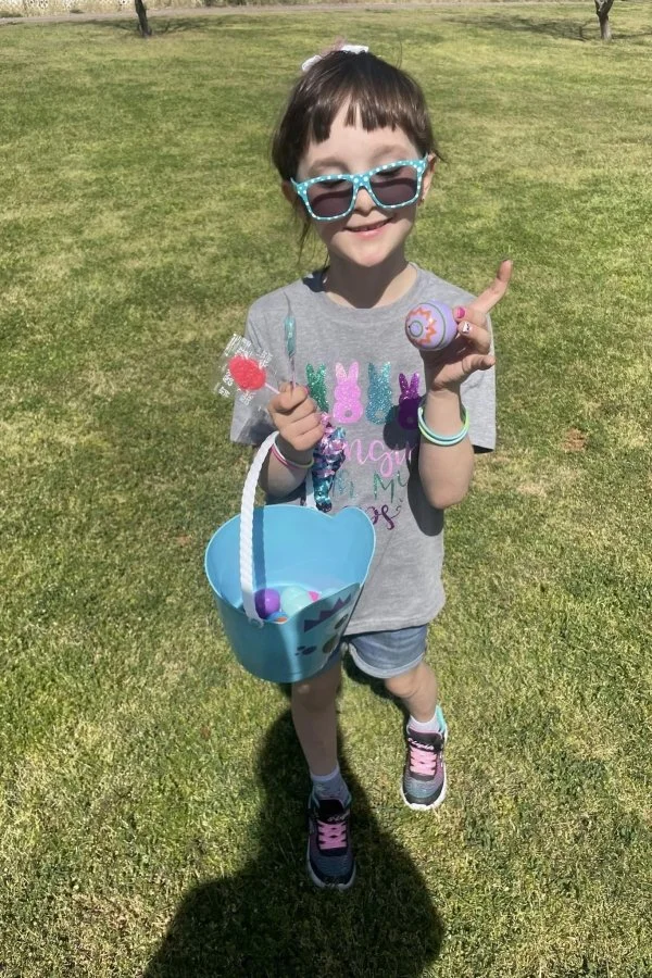 A young girl wearing sunglasses and a gray t-shirt with colorful bunny designs stands on a grassy field. She is holding a small plastic bag with a red candy and a plastic egg, and a blue bucket with purple and pink plastic eggs. The girl has a big smile and appears to be participating in an outdoor Easter egg hunt.
