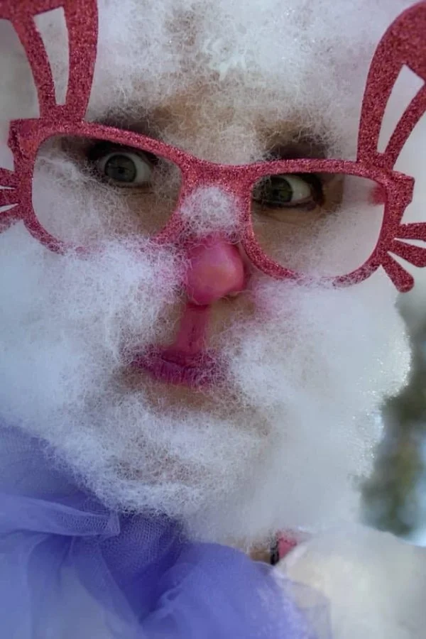 Person in a Santa costume with exaggerated white beard, pink glasses shaped like bunny ears, pink nose, and pink lipstick, close-up.