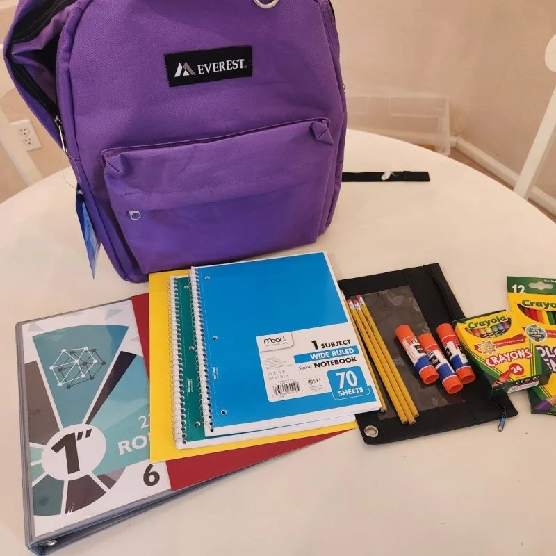A purple Everest backpack on a table with school supplies including notebooks, colored pencils, glue sticks, Crayola crayons, and a binder.