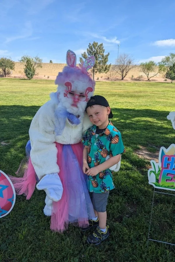 A child smiling and hugging a person dressed as a colorful bunny with pink, purple, and blue fur, wearing glasses, a blue bow tie, and a pastel pink and purple tutu, outdoors on a sunny day.