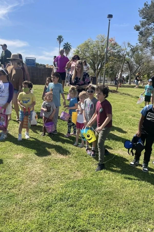 Children holding Easter baskets, standing in a line on a grassy area outdoors on a sunny day, likely participating in an Easter egg hunt.