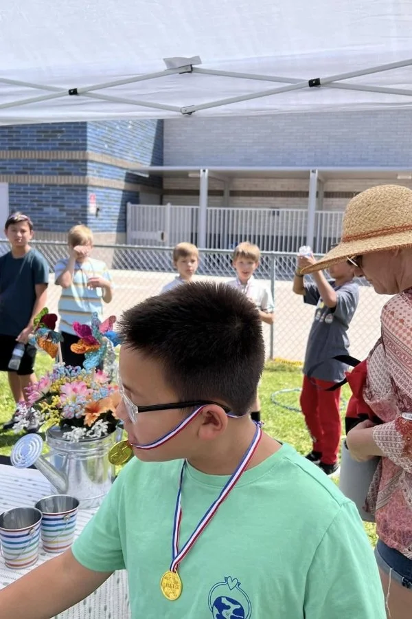 A boy wearing glasses and medals around his neck is at an outdoor event, tasting from a table with cups and colorful flower arrangements. Other children and an adult wearing a straw hat are seen in the background near a fence and a building.