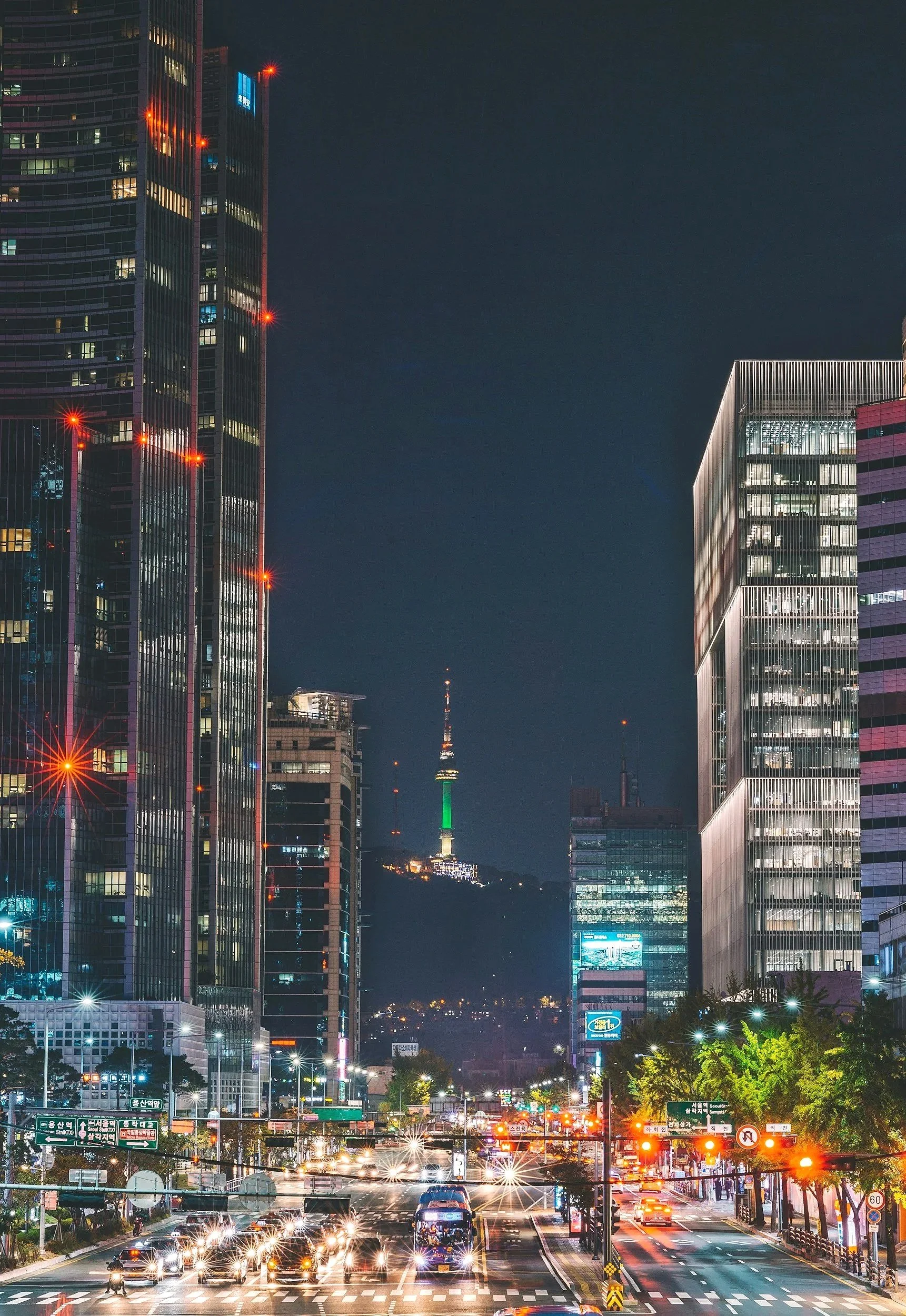 Night cityscape with tall illuminated skyscrapers, a busy traffic street with cars and buses, and a tall tower with colorful lights in the distance.