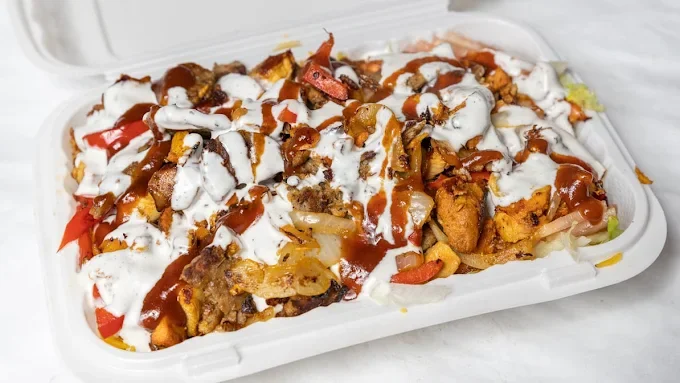 Takeout container filled with nachos topped with sour cream, hot sauce, and chopped vegetables.