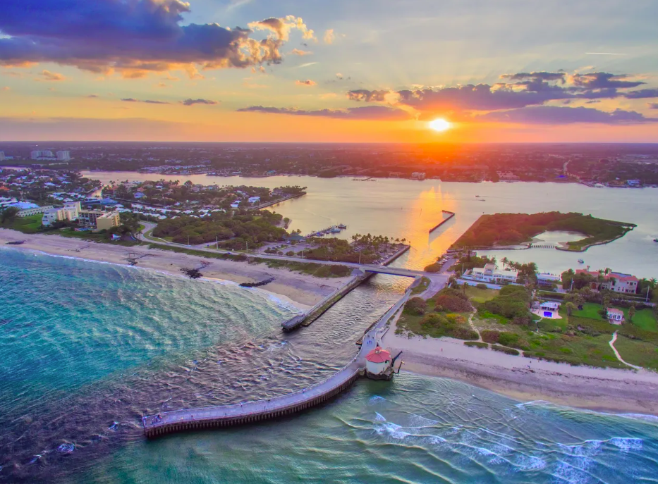 Aerial view of a coastal area at sunset with a pier extending into the ocean, sandy beaches, calm waters, residential buildings, and greenery along the shoreline.