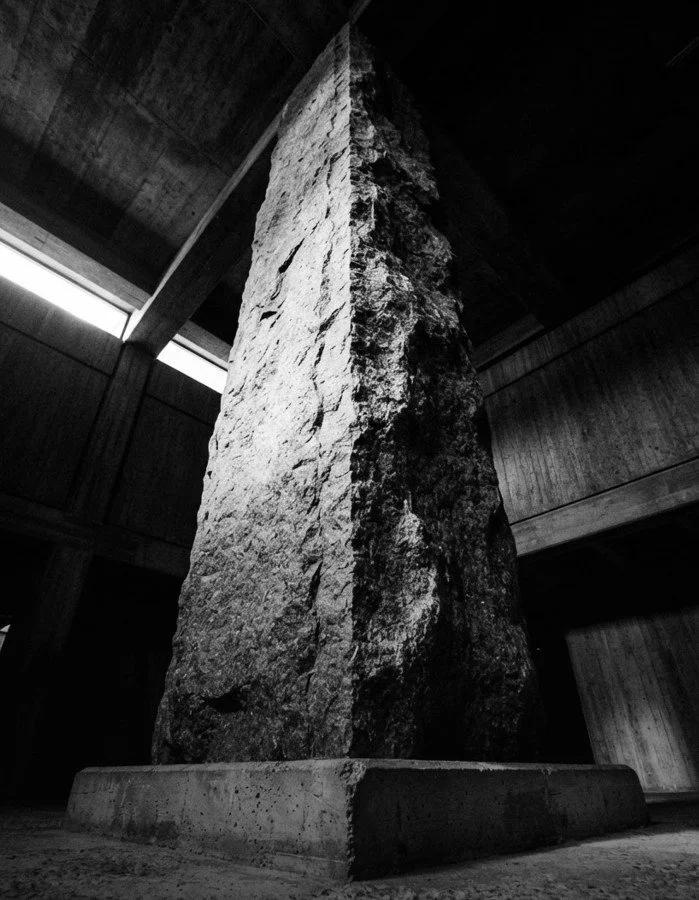 A tall, rough-textured stone monument standing on a concrete base inside a wooden structure with high ceilings.