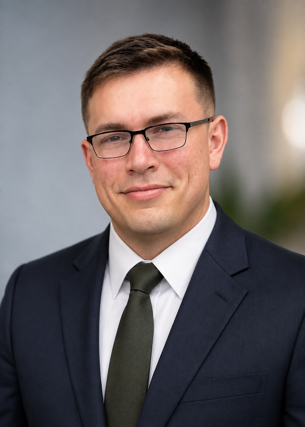 Headshot of a young man in a dark suit, white shirt, and dark tie, wearing glasses, smiling against a blurred neutral background.