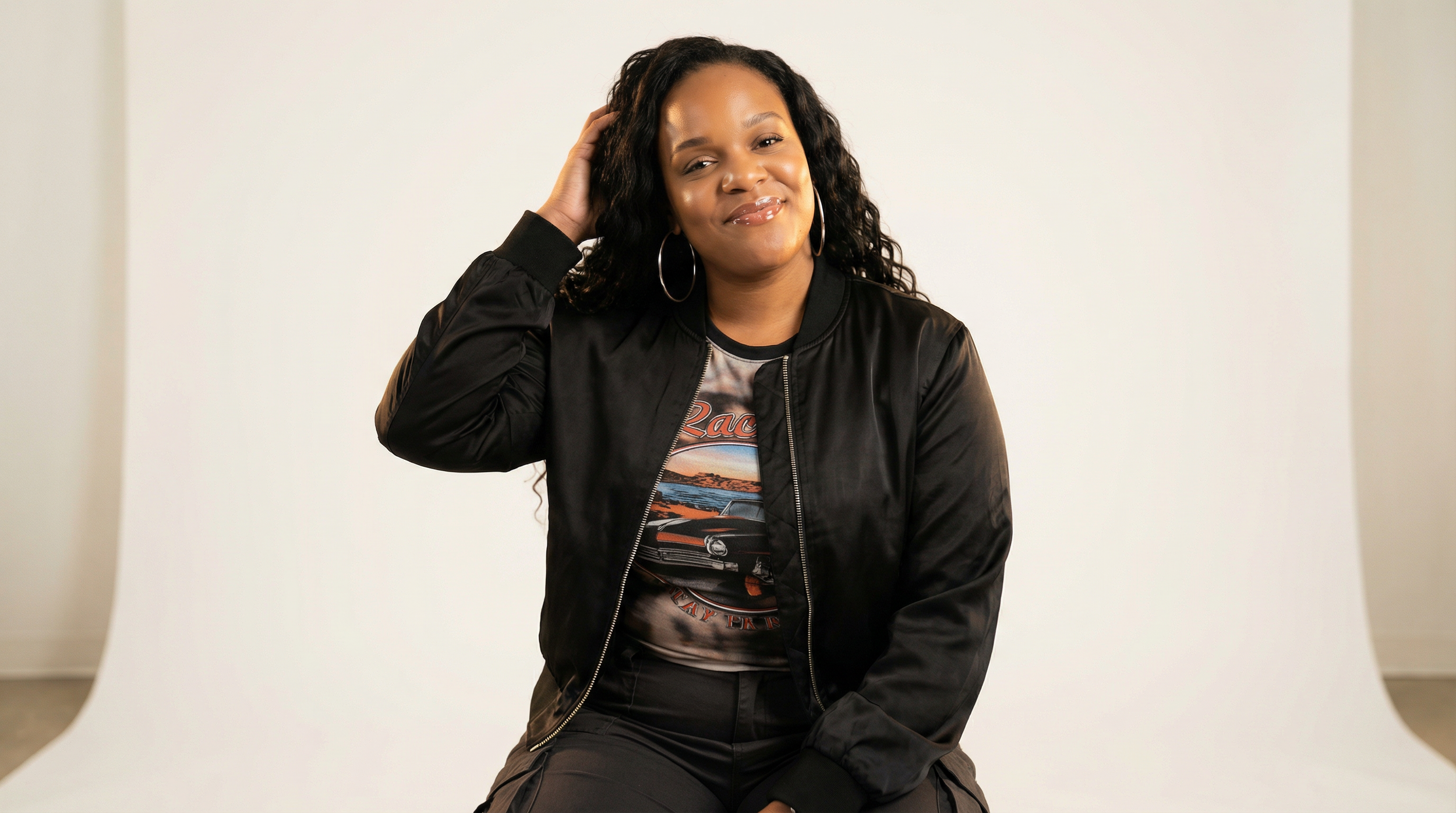 A woman with long curly black hair wearing hoop earrings, a black jacket, and a graphic t-shirt, sitting against a white background, smiling with her hand touching her head.
