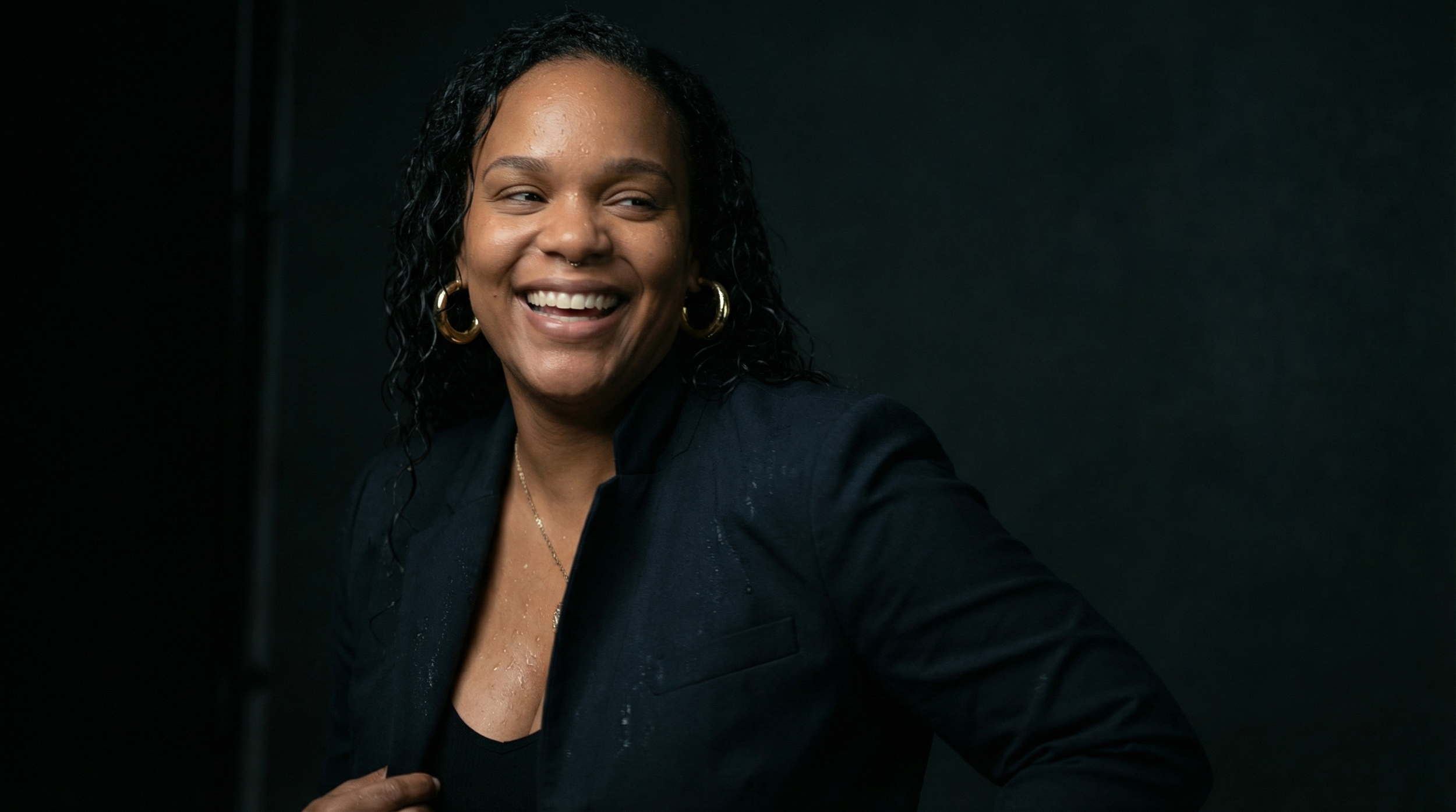A woman with curly black hair, gold hoop earrings, and a black blazer smiling in front of a dark background.