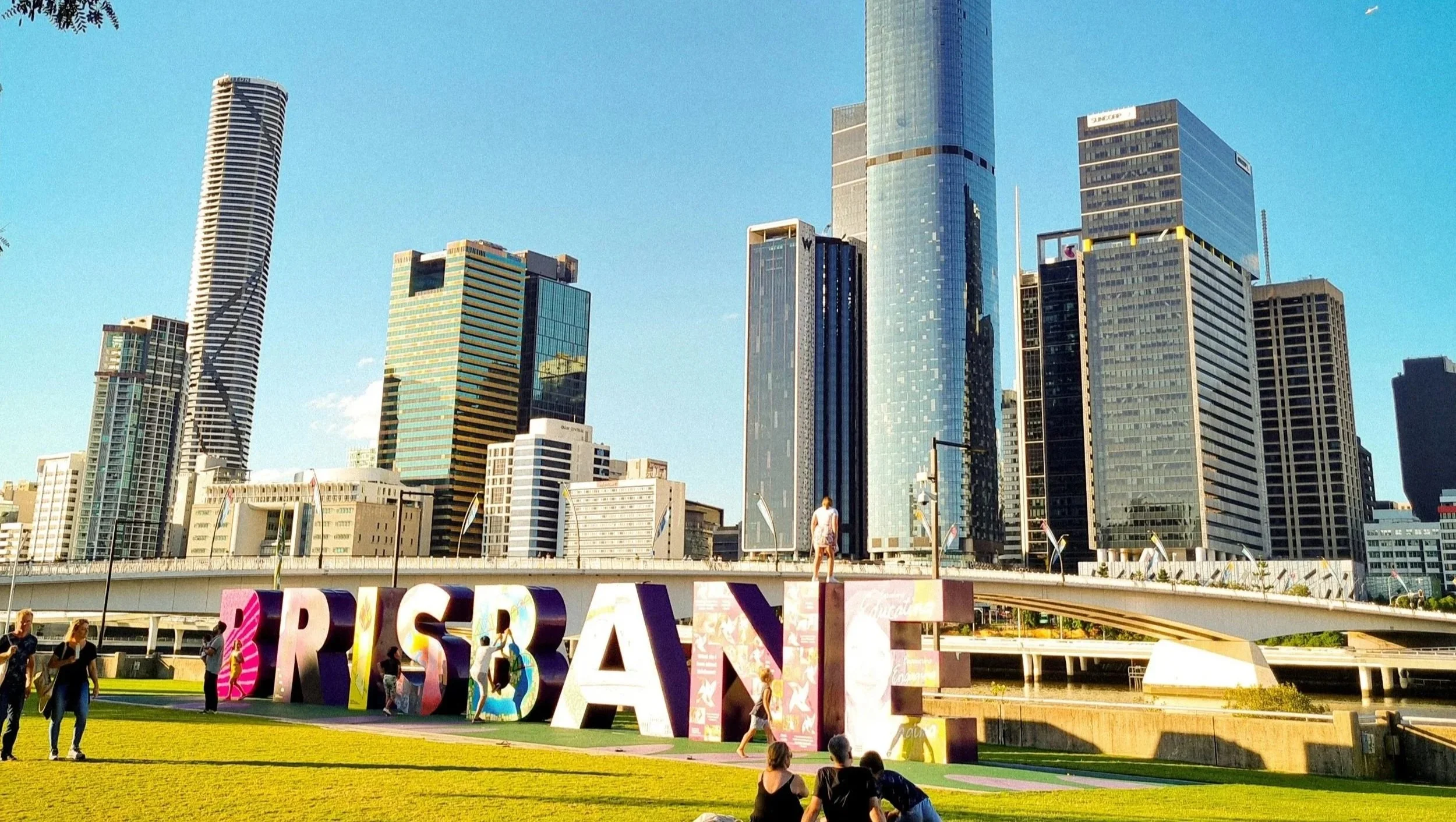 People gathered in a park with large colorful 'BRISBANE' letters in front of a city skyline with modern high-rise buildings.