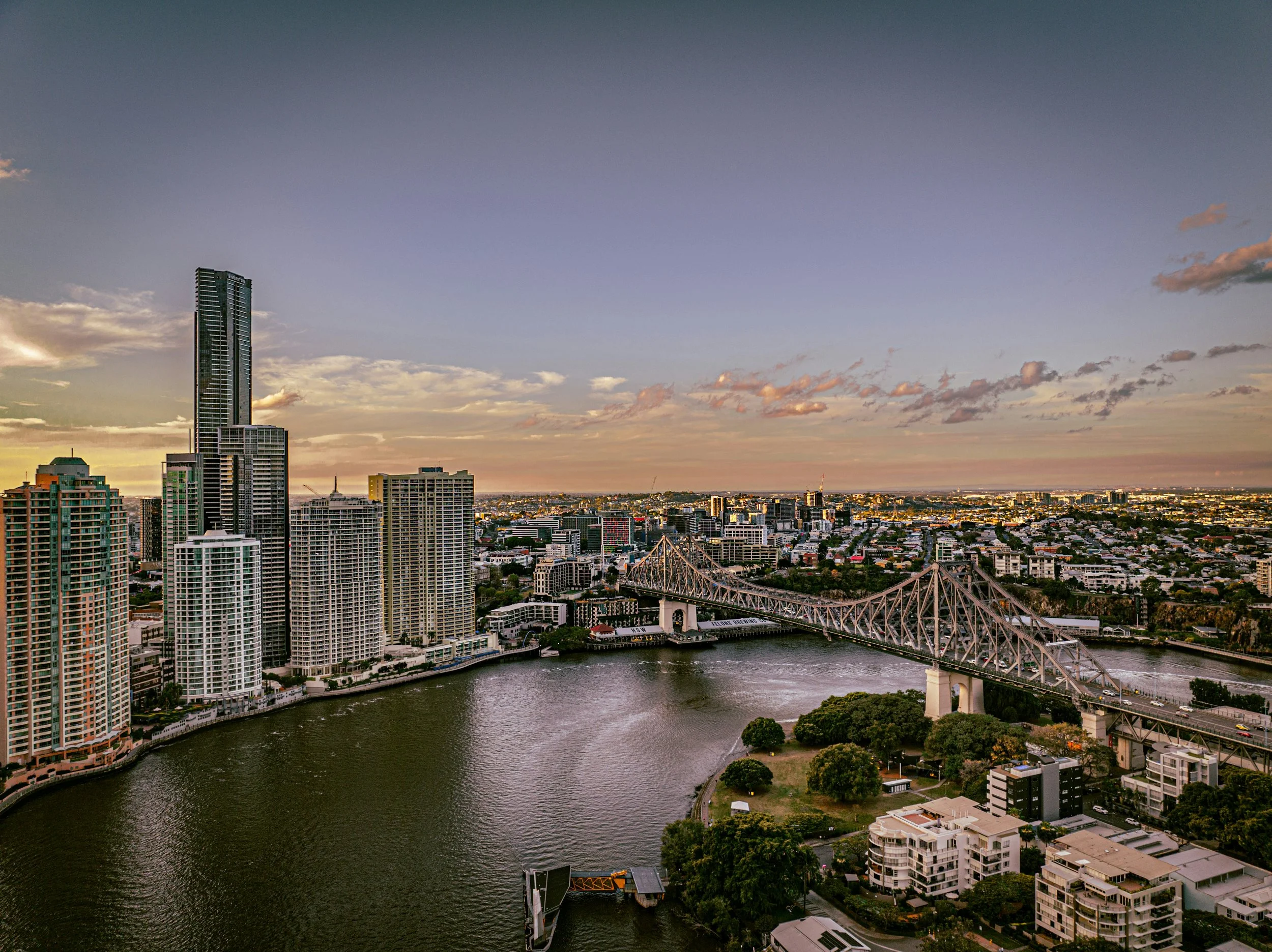 Skyline of a city during sunset with high-rise buildings, a river flowing through the city, and a bridge over the water, with a mostly clear sky and a few clouds.