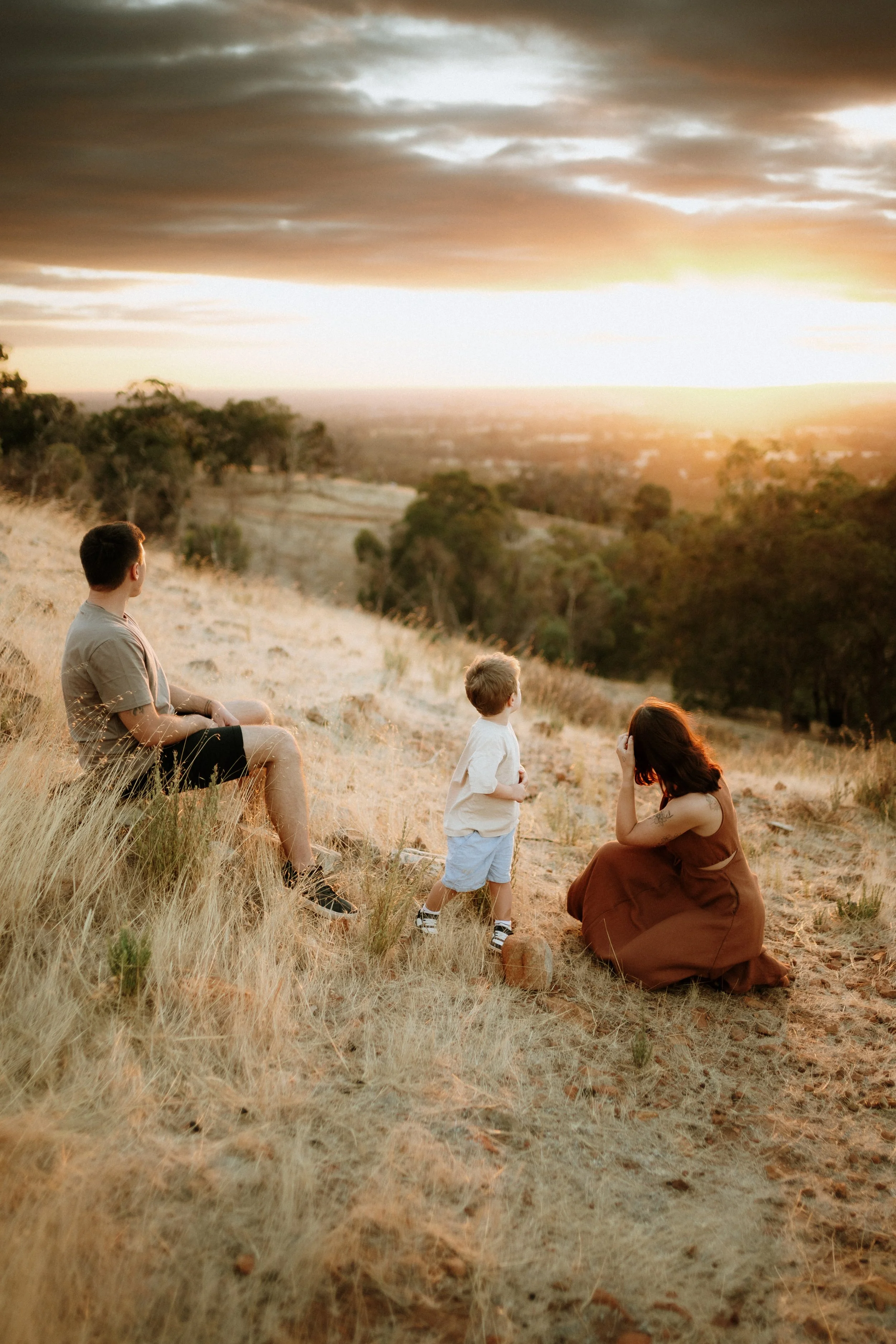 A group of people, including a man, a woman, and two children, sitting and standing on a hillside covered with dry grass, watching a sunset or sunrise with clouded sky.