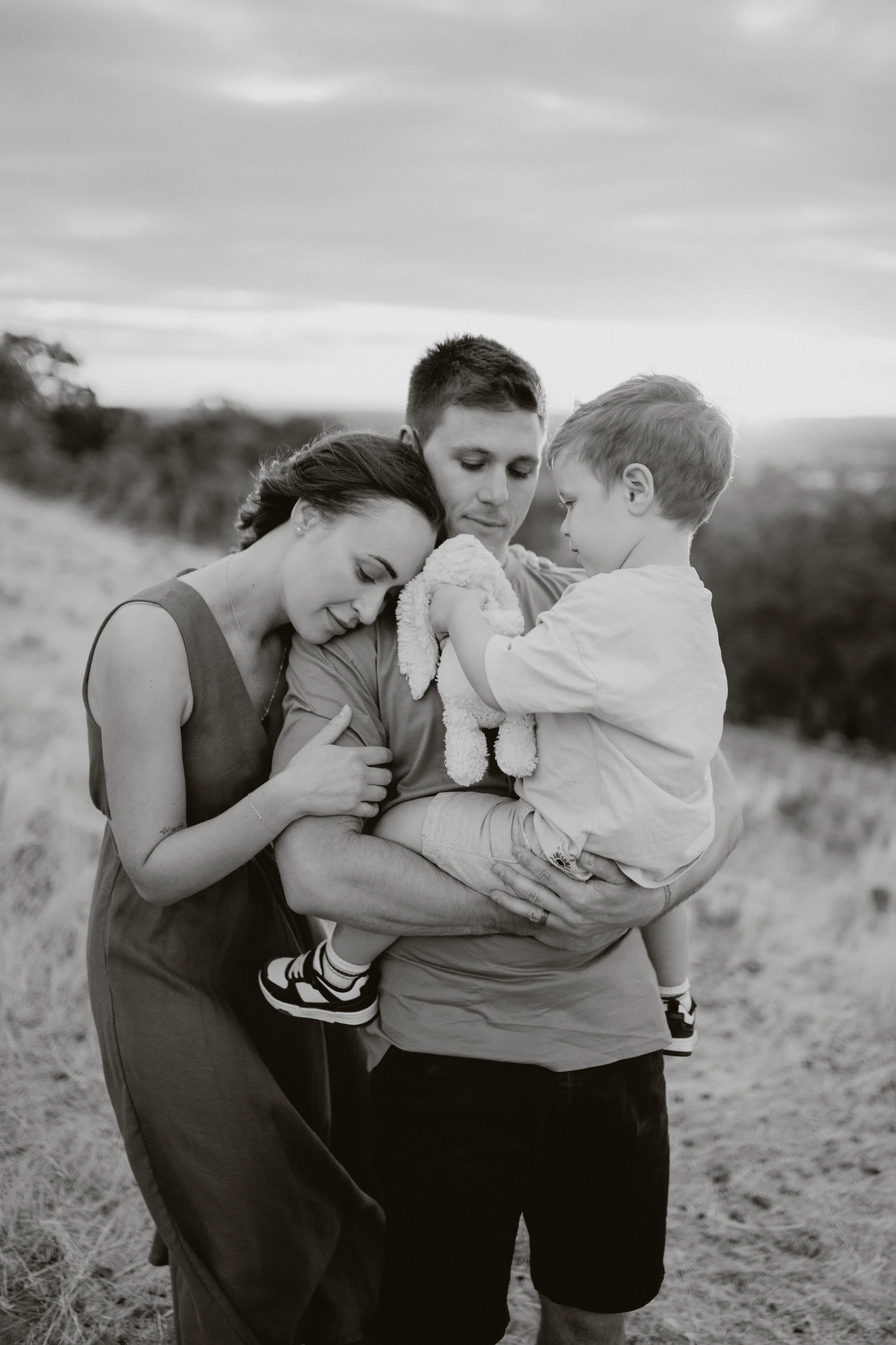 A family of four, including a woman, man, and two young children, sharing a hug outdoors on a cloudy day.