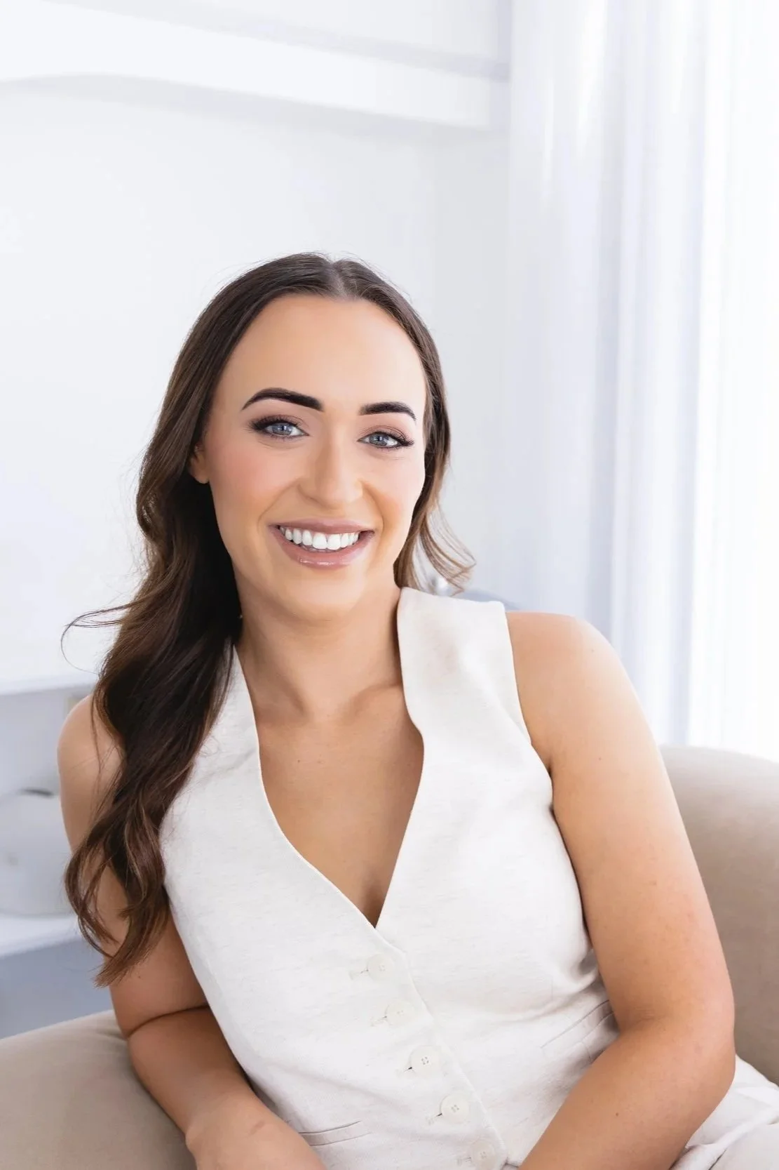 Smiling woman with long brown hair and blue eyes, wearing a sleeveless white top, sitting on a light-colored couch in a bright, modern room.
