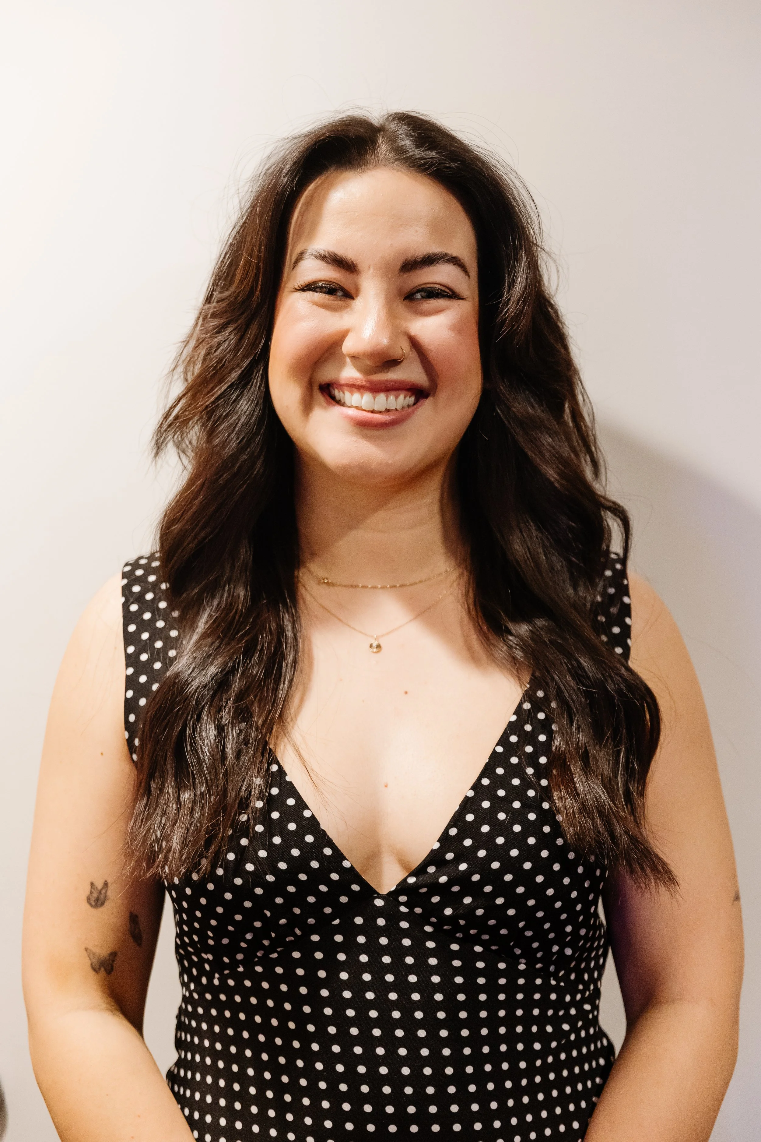 A smiling woman with long, wavy dark hair, wearing a black polka-dot sleeveless dress, gold necklaces, and small tattoos of butterflies on her left arm, standing against a plain, light-colored wall.
