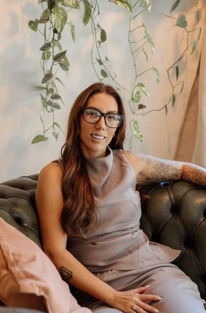 A woman with long brown hair, wearing glasses and a beige sleeveless dress, sitting on a dark green tufted leather sofa with patterned pillows, in front of a light-colored wall with hanging green vines.