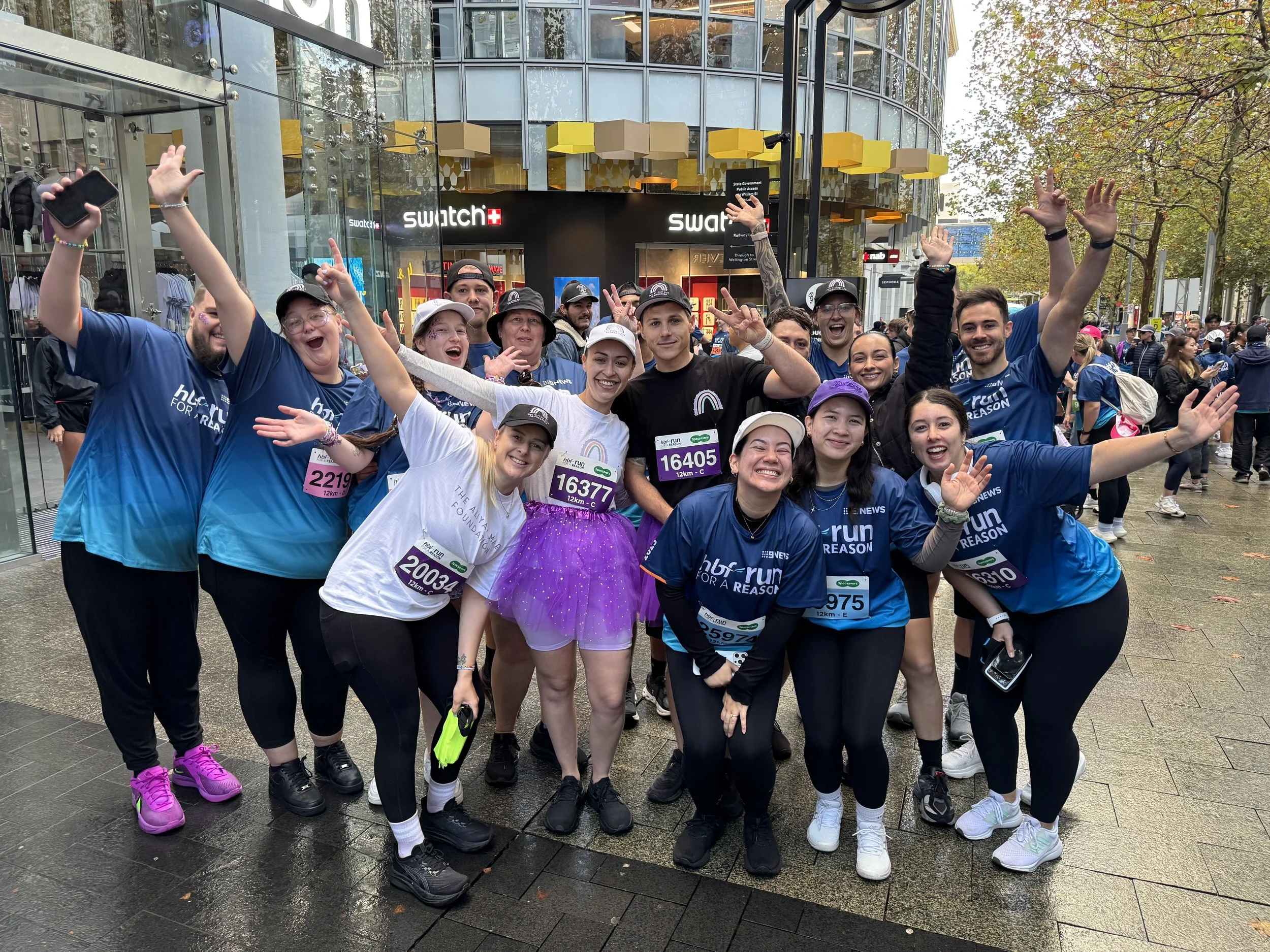 A group of people in athletic clothing and race bibs gathered in a city street, smiling and celebrating after participating in a running event, with a modern glass building and crowded street in the background.