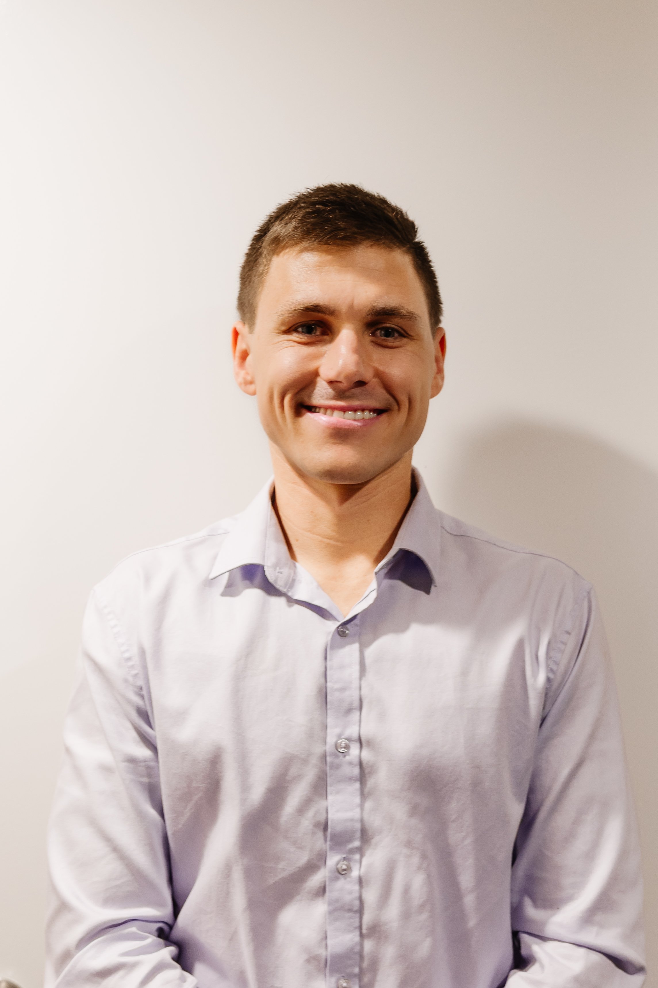 A young man with short brown hair wearing a light purple button-up shirt, smiling, standing against a plain off-white wall.
