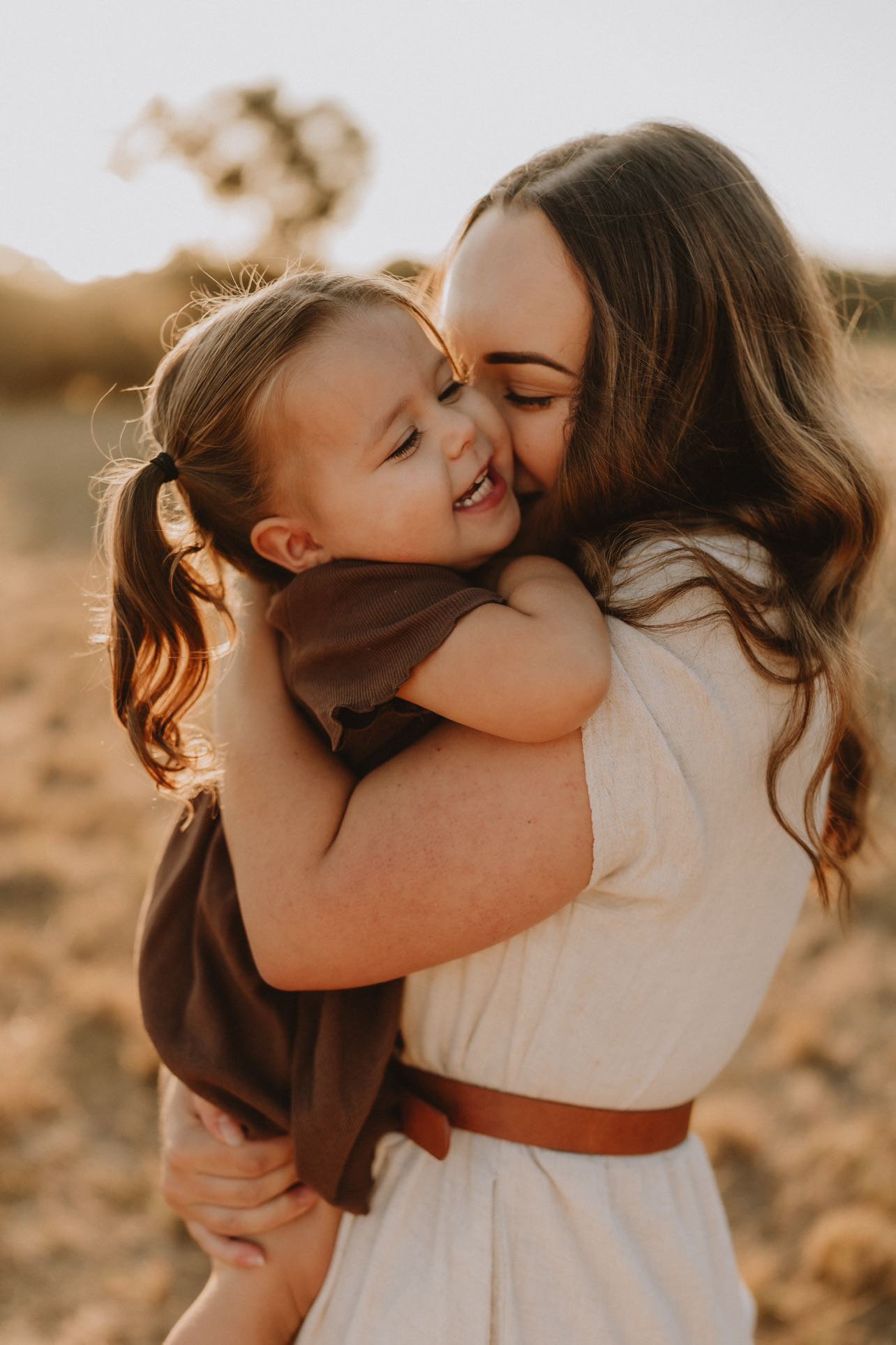 A woman holding a young girl in a tight embrace outdoors at sunset, smiling and laughing.