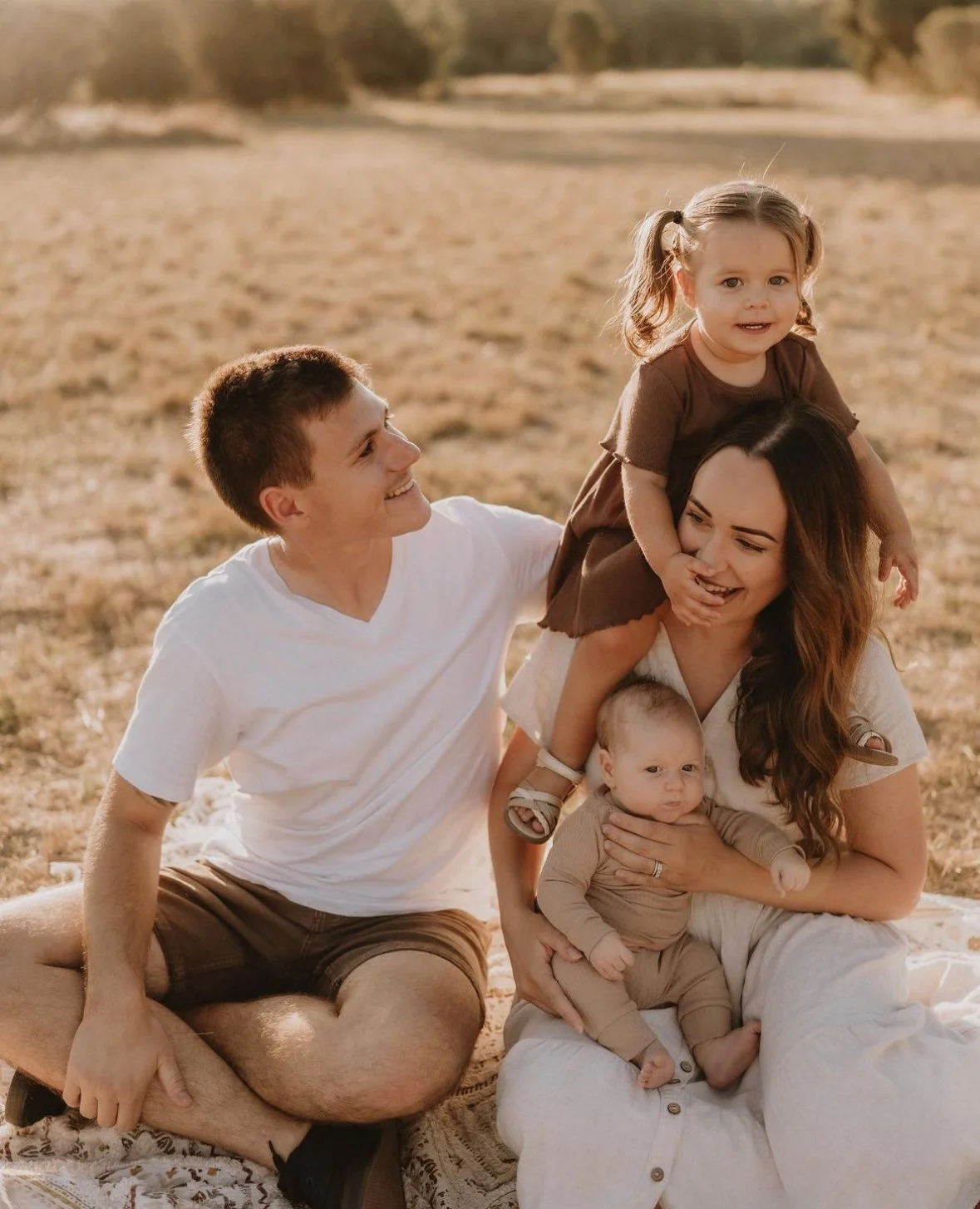 A family of four with two young children enjoying time outdoors on a sunny day, sitting on a blanket in a grassy field.