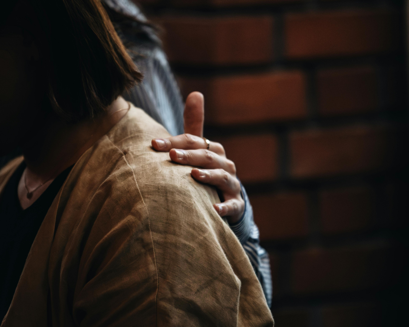 Close-up of a person's hand resting on another person's shoulder, with a brick wall in the background.