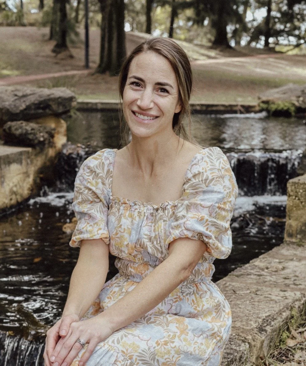 A woman with brown hair sitting on stone beside a small waterfall or stream in a park, smiling at the camera.