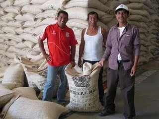 Three people standing in front of large sacks of rice or flour inside a warehouse.