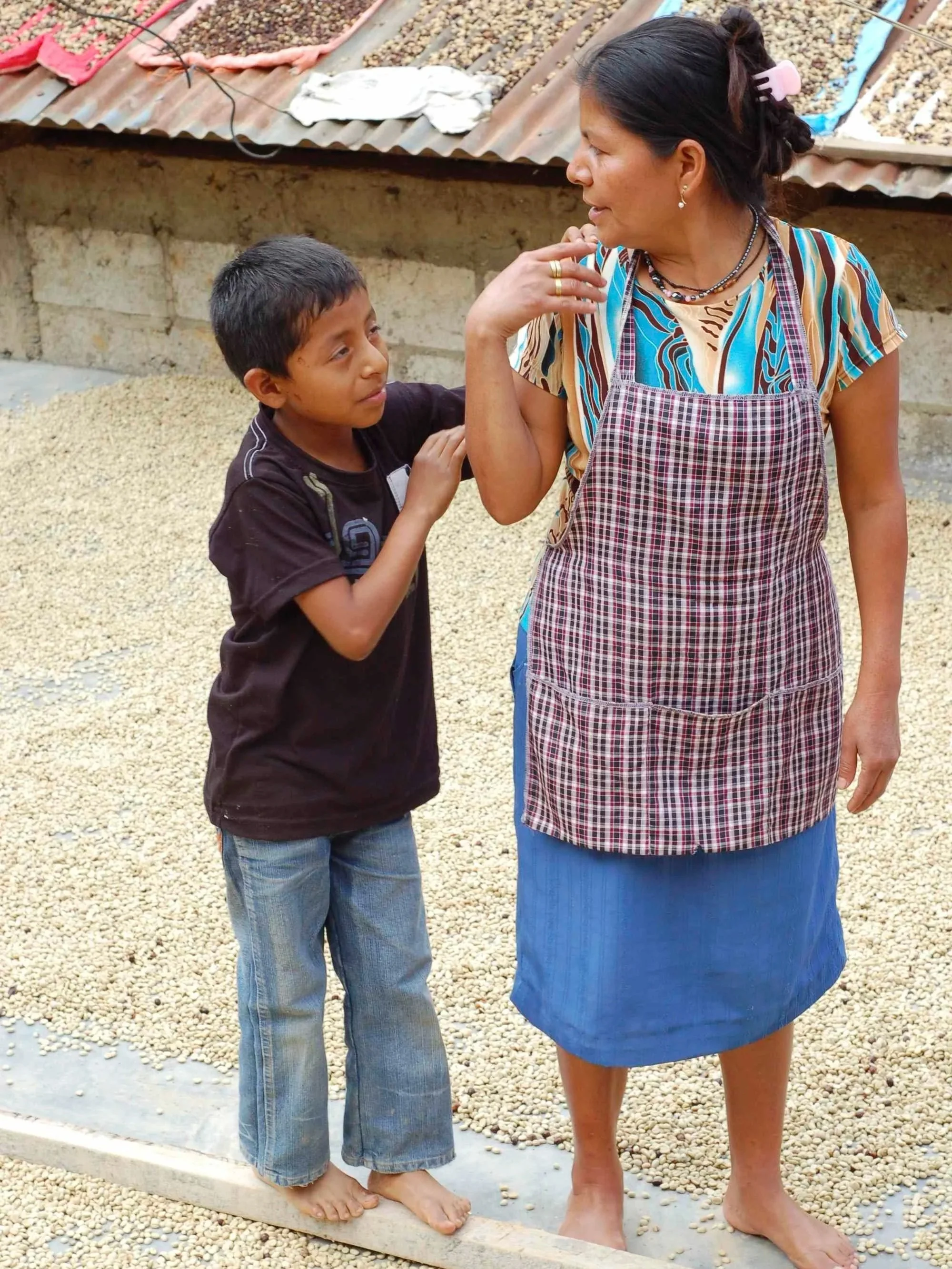 A woman and a young boy standing on a gravel surface, engaged in conversation. The woman is wearing a patterned dress with a checkered apron and has her hair tied back with a pink clip. The boy is barefoot, wearing a black t-shirt and blue jeans, and