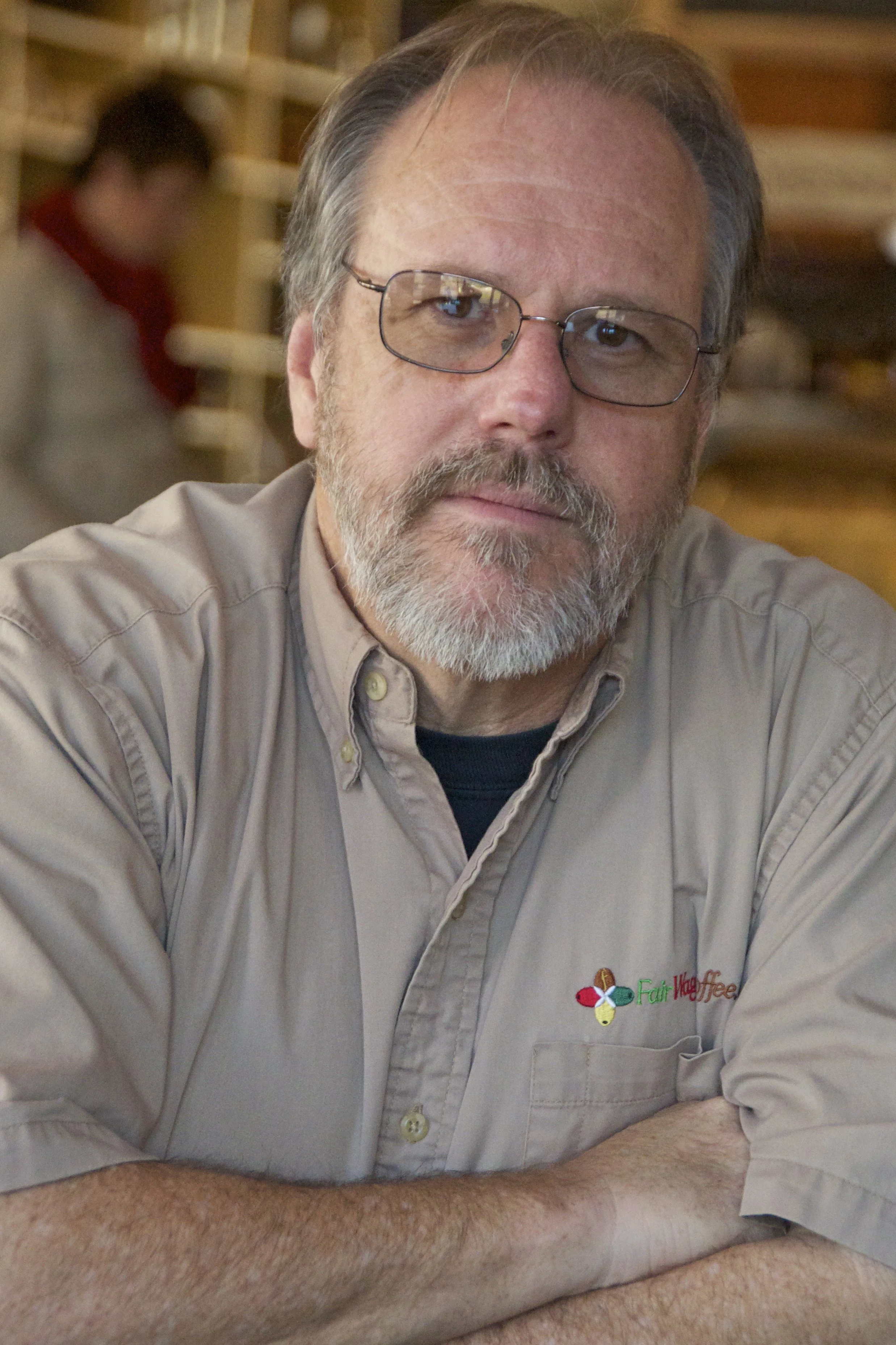 A middle-aged man with glasses and a beard wearing a beige shirt with a 'FairVkeffee' logo, sitting at a table, arms crossed, in a cozy indoor setting with another person blurred in the background.