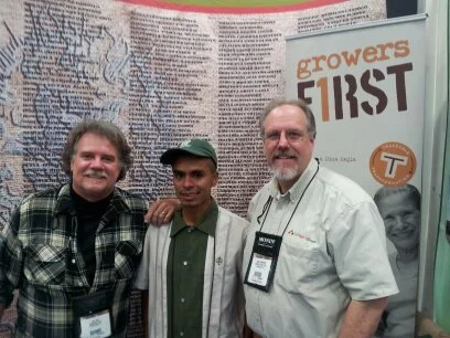 Three men standing together at an indoor event, with a large informational poster and a banner that reads 'growers FIRST' visible in the background.