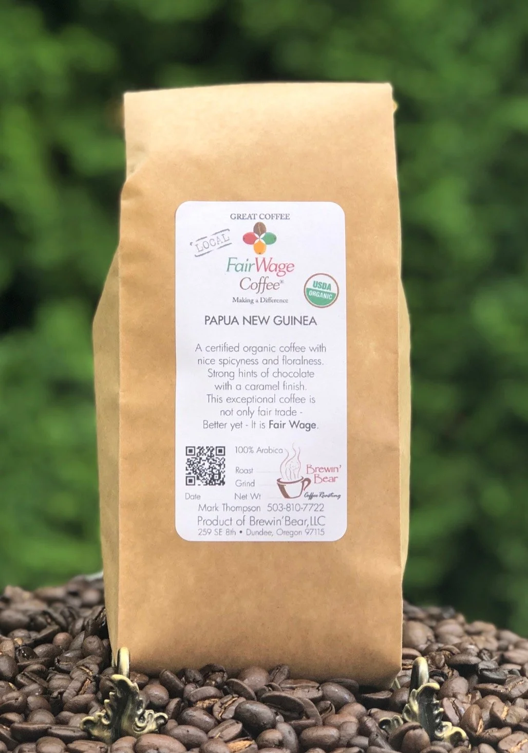 Bag of Fair Wage Coffee from Papua New Guinea, with a white label showing organic certification, QR code, and branding, set on a bed of coffee beans.