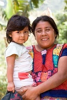 A woman holding a young girl outdoors, both smiling, with greenery in the background.