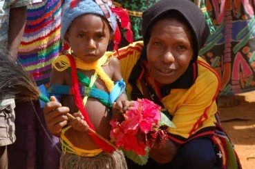 A young girl with traditional attire holding a bouquet of pink flowers, standing next to an adult woman in a yellow shirt, both outdoors among other people.