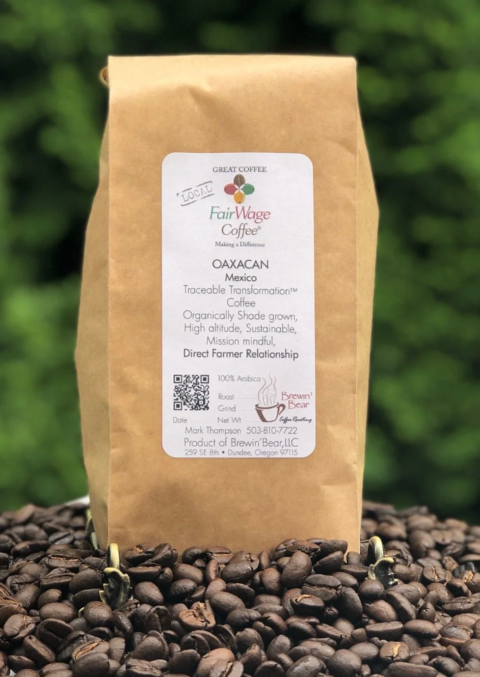 A brown paper bag of FairWage Coffee, labeled as OAXACAN from Mexico, placed on a bed of dark roasted coffee beans with a blurred green background. The label includes a QR code, contact information, and describes the coffee as traceable, organically 