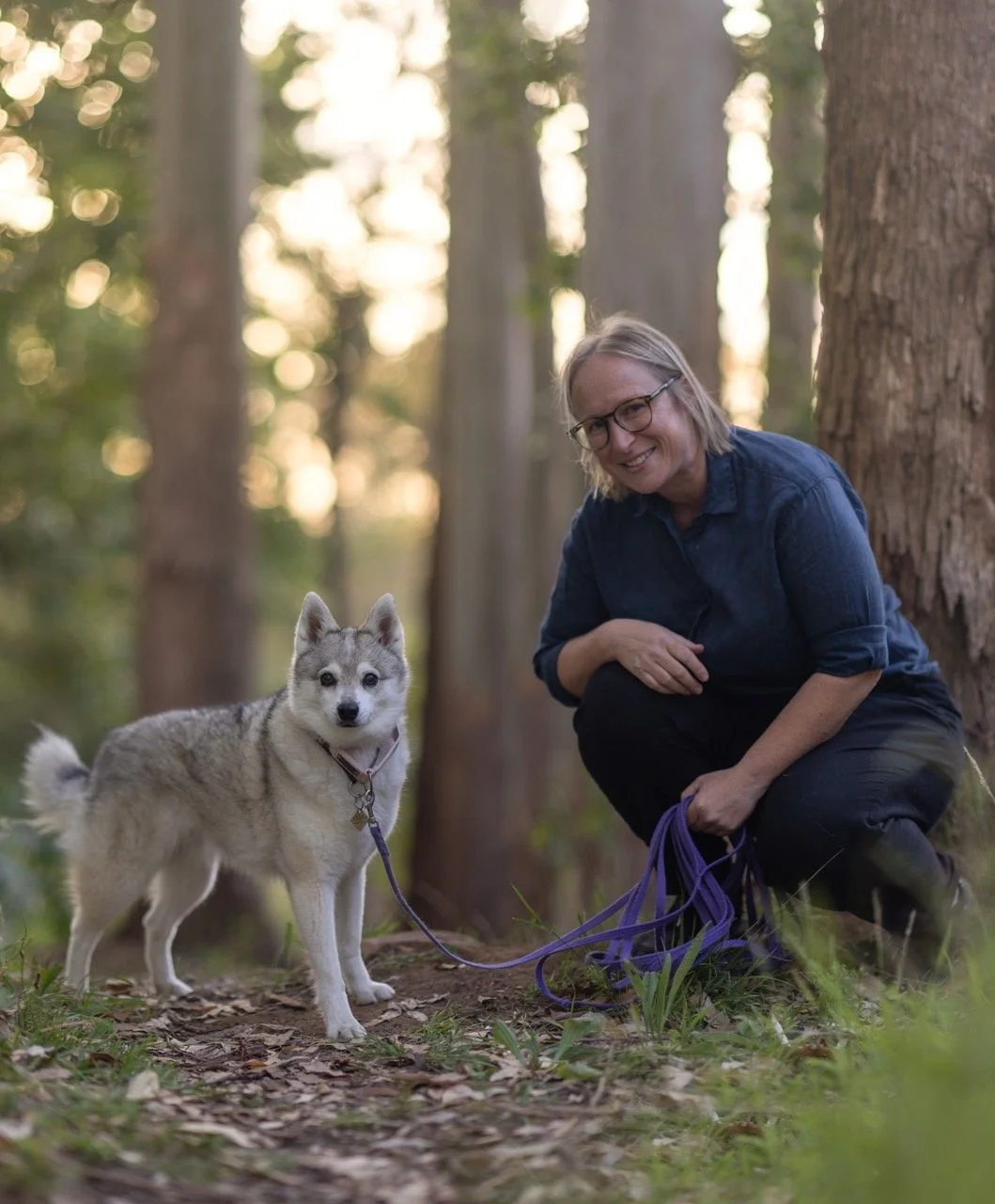 A woman with short hair and glasses crouches next to an alaskan klee kai dog on a leash in a forest, with tall trees and blurred green foliage in the background.