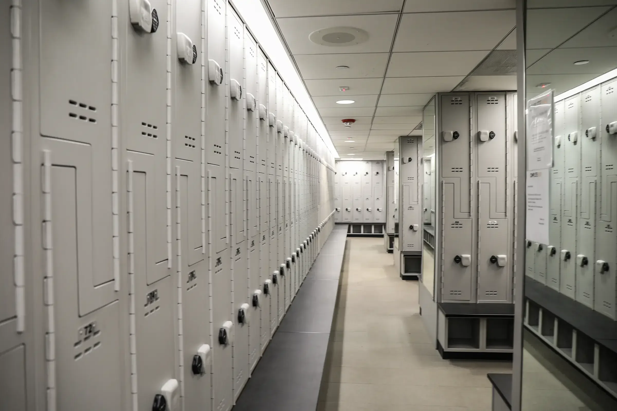 Row of white lockers in a hallway of a school or gym, some with combination locks, with ceiling lights and a fire sprinkler system.