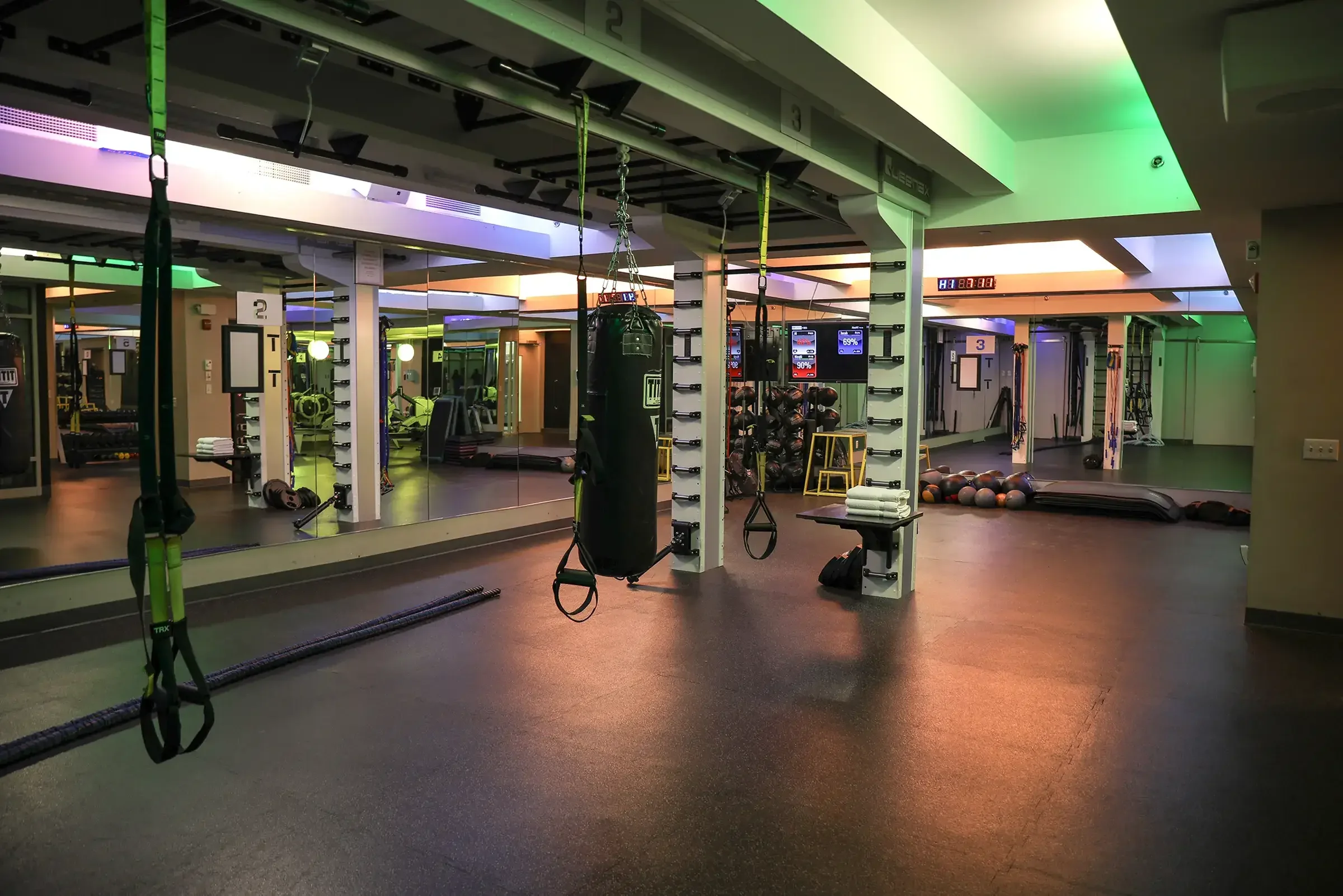 Empty fitness gym with workout equipment, mirrored walls, and colorful ceiling lighting.