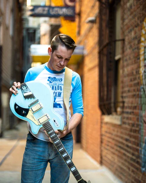 A man holding a light blue electric guitar in a narrow alleyway with brick walls.