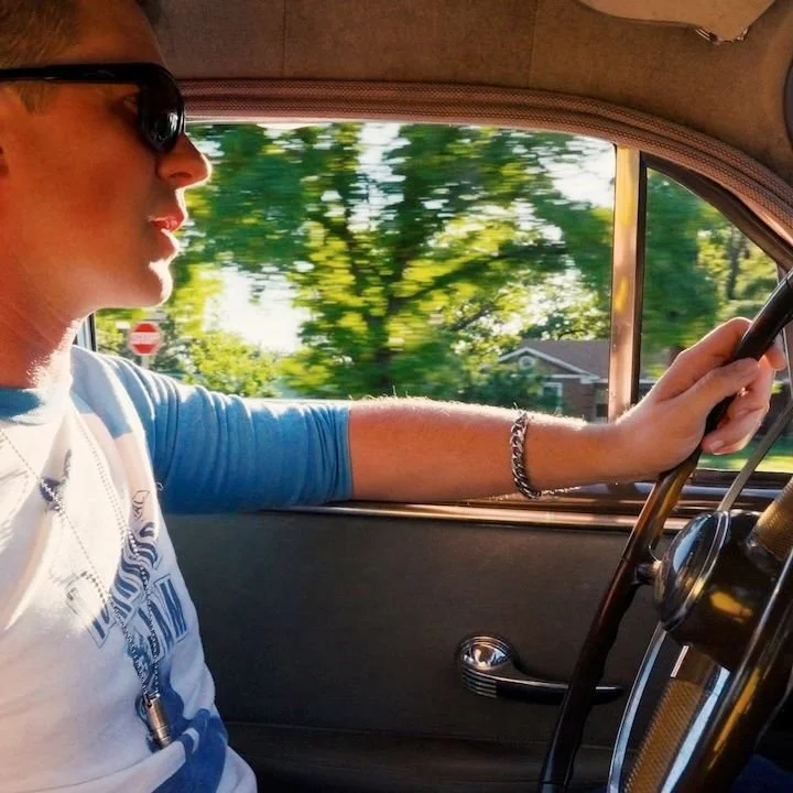 A man wearing sunglasses and a white and blue shirt is driving a vintage car with a gray interior, with greenery and houses visible outside the window.