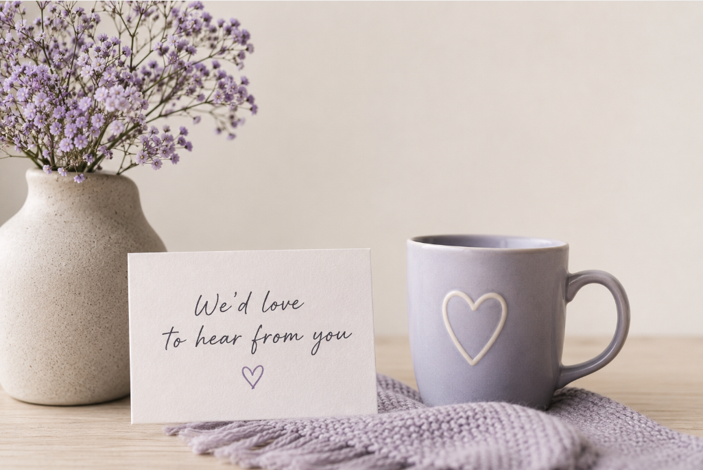 A white vase with purple flowers, a handwritten note saying "We'd love to hear from you" with a small purple heart, a lavender mug with a white heart outline, resting on a lavender knitted cloth on a wooden surface.