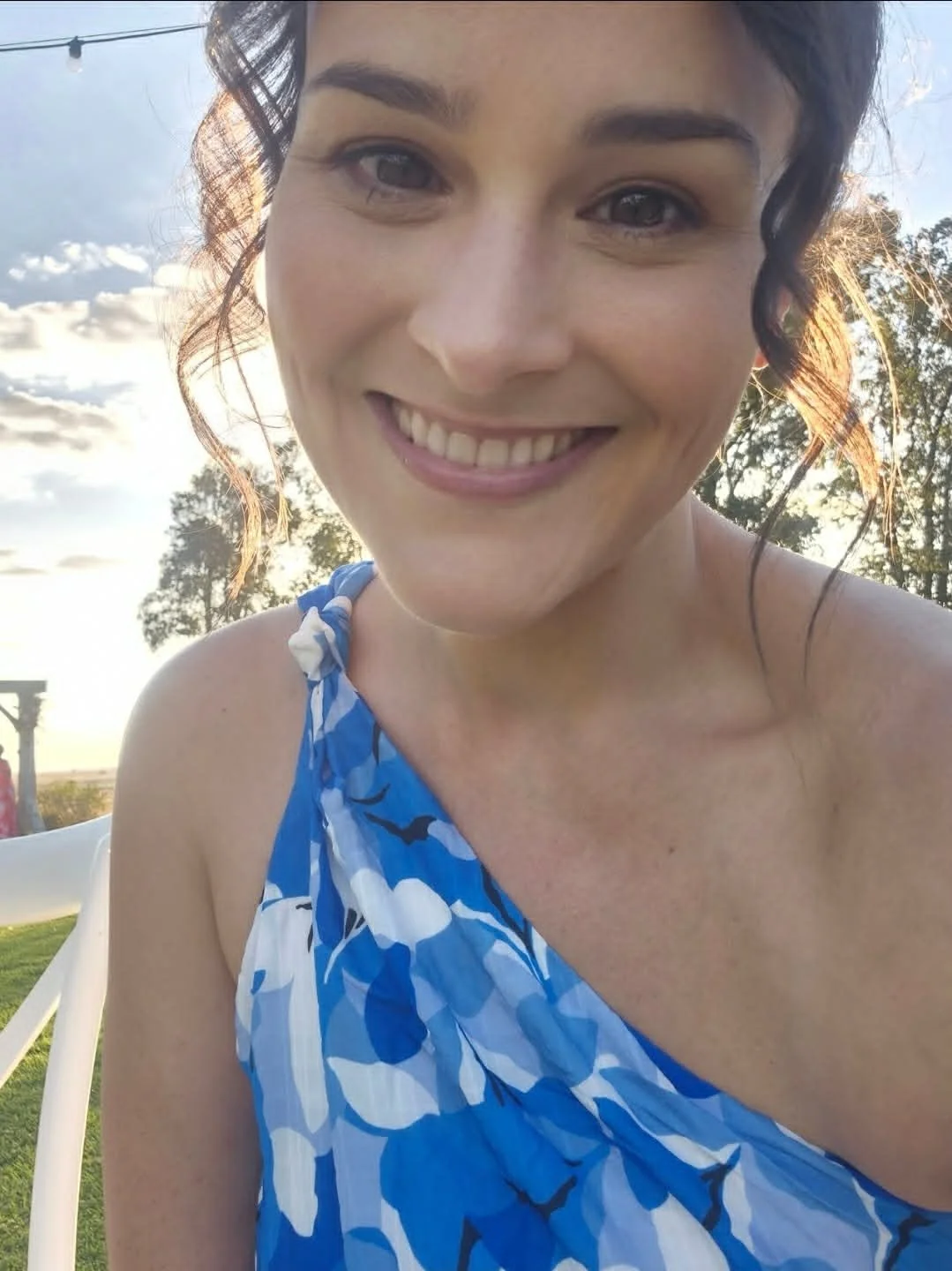 A woman smiling outdoors in a blue and white patterned sleeveless dress during sunset, with trees and a partly cloudy sky in the background.