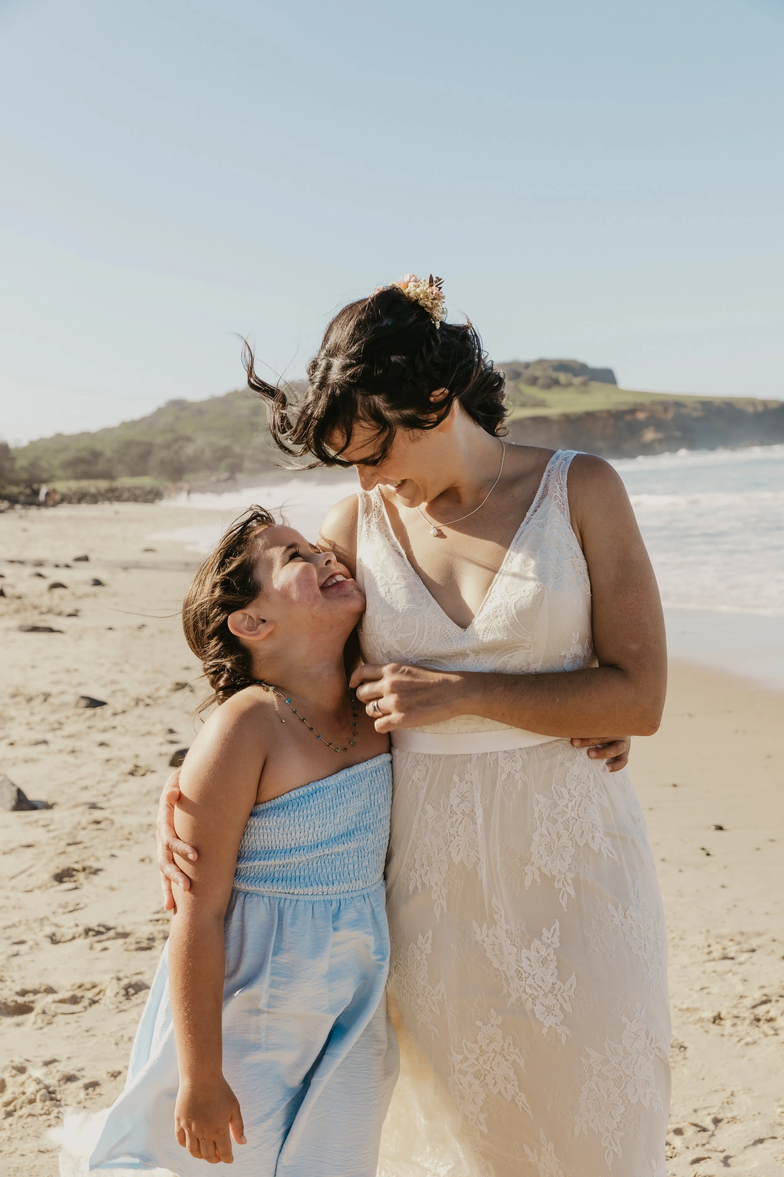 A woman and a young girl are on a sandy beach, smiling and embracing. The woman is wearing a cream lace dress and the girl is in a light blue dress. They are looking at each other happily, with ocean waves and distant land formations in the background.