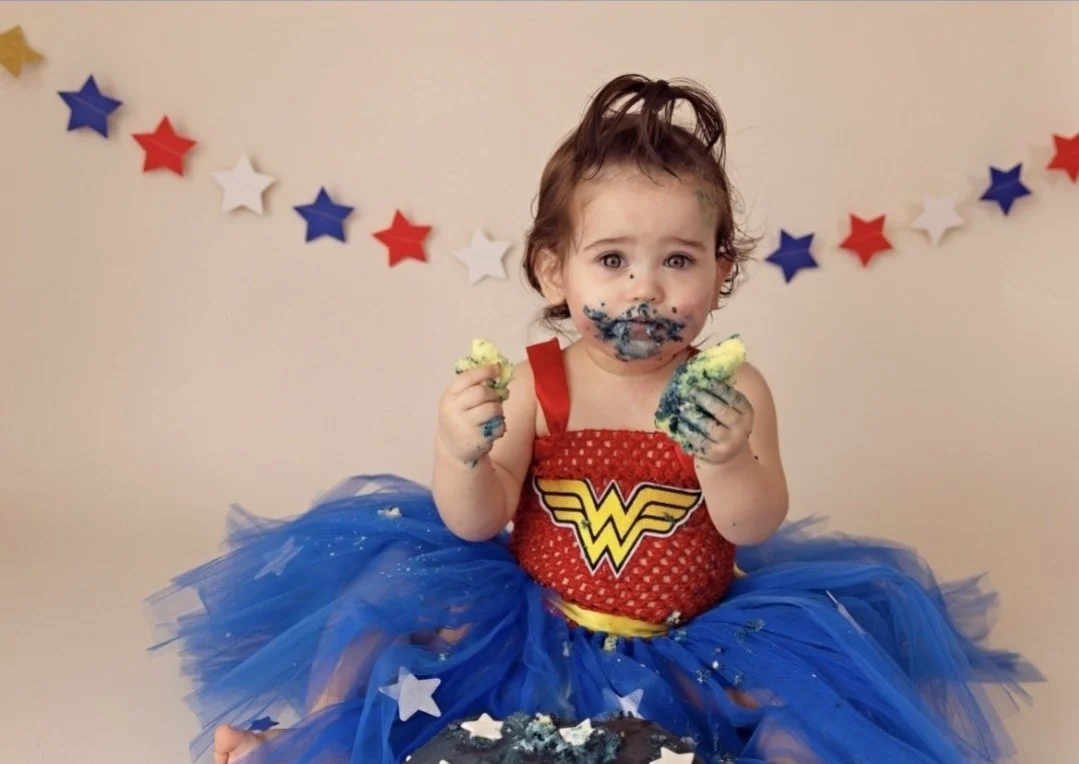 A young girl dressed as Wonder Woman, wearing a blue tutu, sitting with her face and hands covered in blue, yellow, and green frosting, celebrating her birthday.